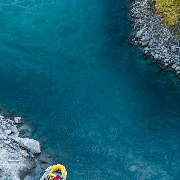 looking down on river from up high showing water flow yellow dingy and two men lying in boat with rocks on river shore and small jetty and green bushes in queenstown bungy jump area